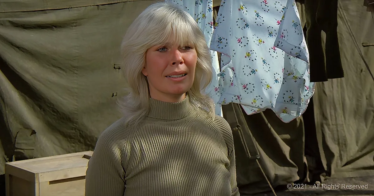 A woman with blonde hair in a turtleneck sweater stands in front of a tent with clothes hanging.
