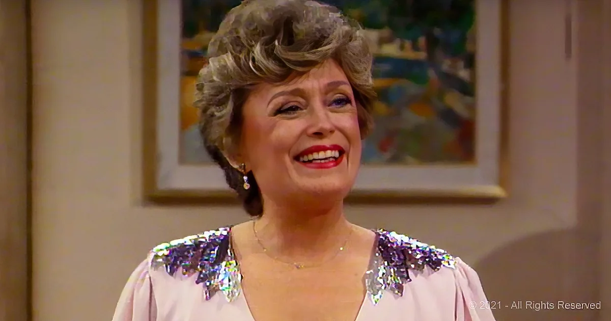 A smiling woman with short, curly hair and a sparkly shoulder detail in a light-colored top.