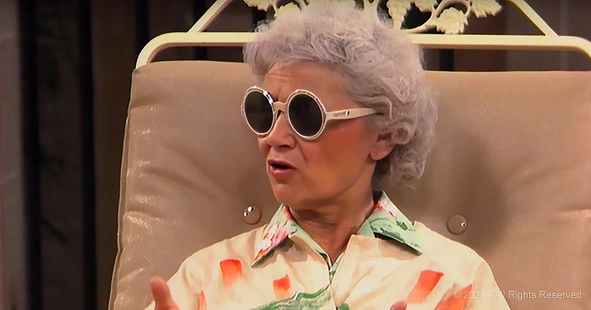 An elderly woman with gray hair and sunglasses sits in a chair, wearing a floral shirt, and appears to be speaking.