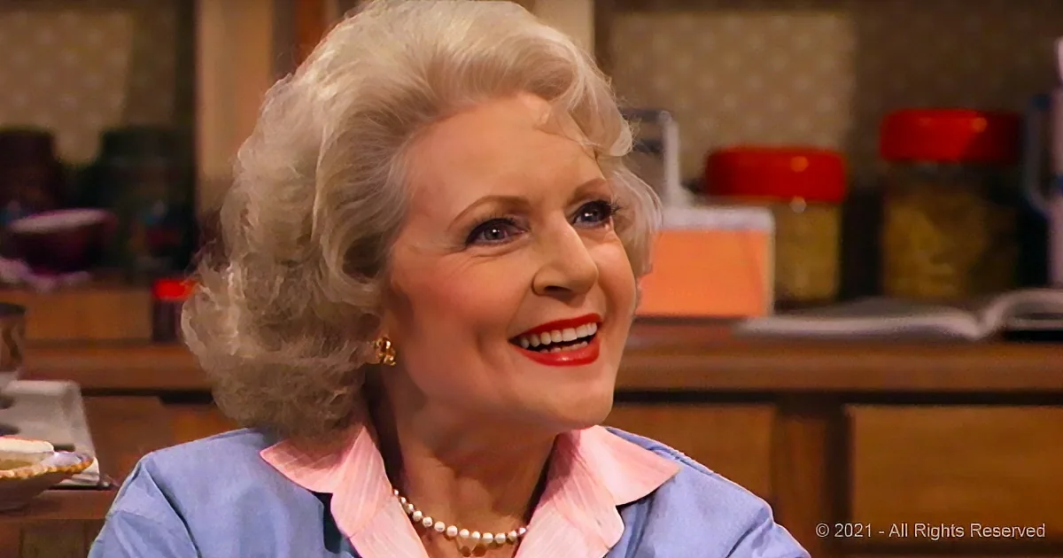 A smiling woman with gray hair, wearing a blue blouse and pearl necklace, in a kitchen setting.