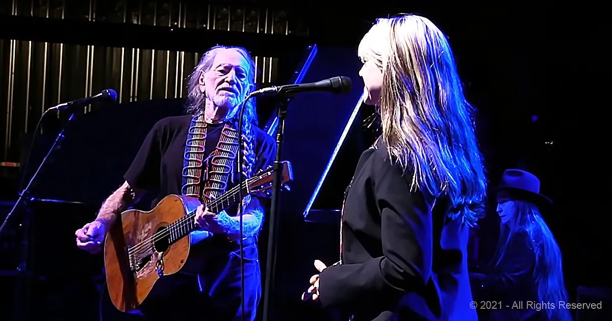 A man with long hair and beard plays guitar while a woman sings into a microphone on stage.