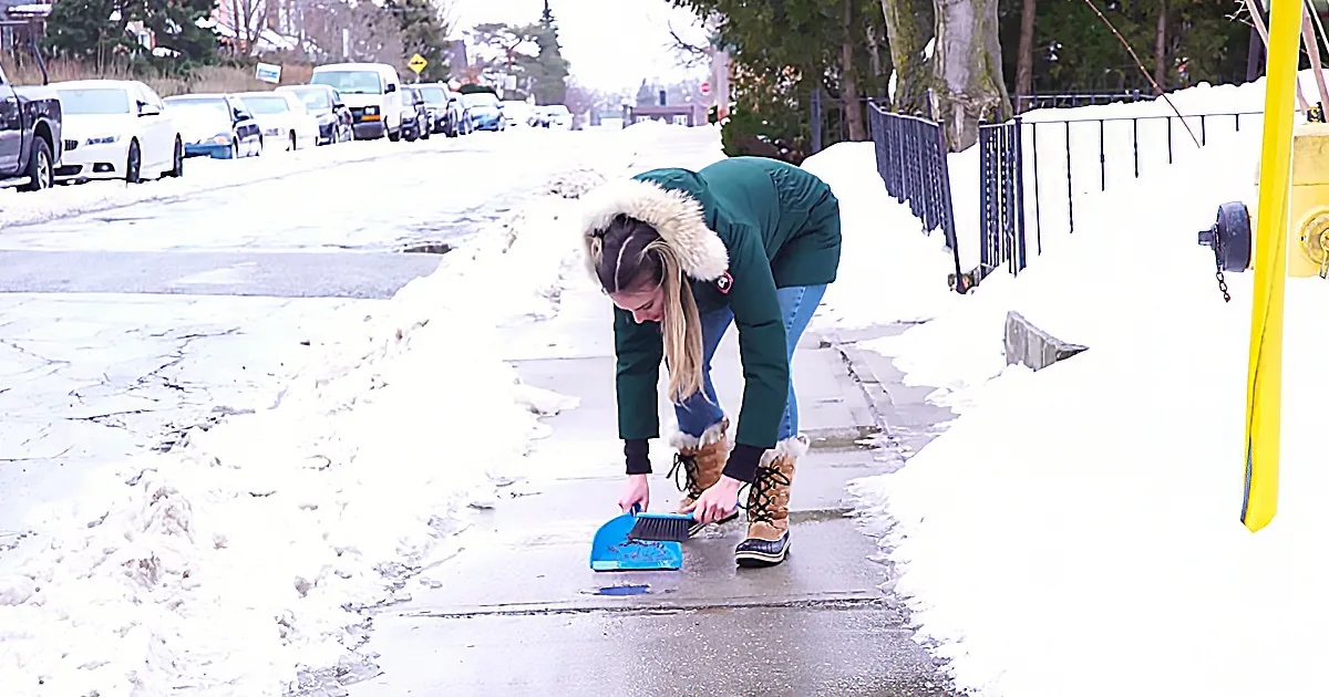 A woman in a green coat and brown boots is sweeping a wet sidewalk near a snow-covered street.