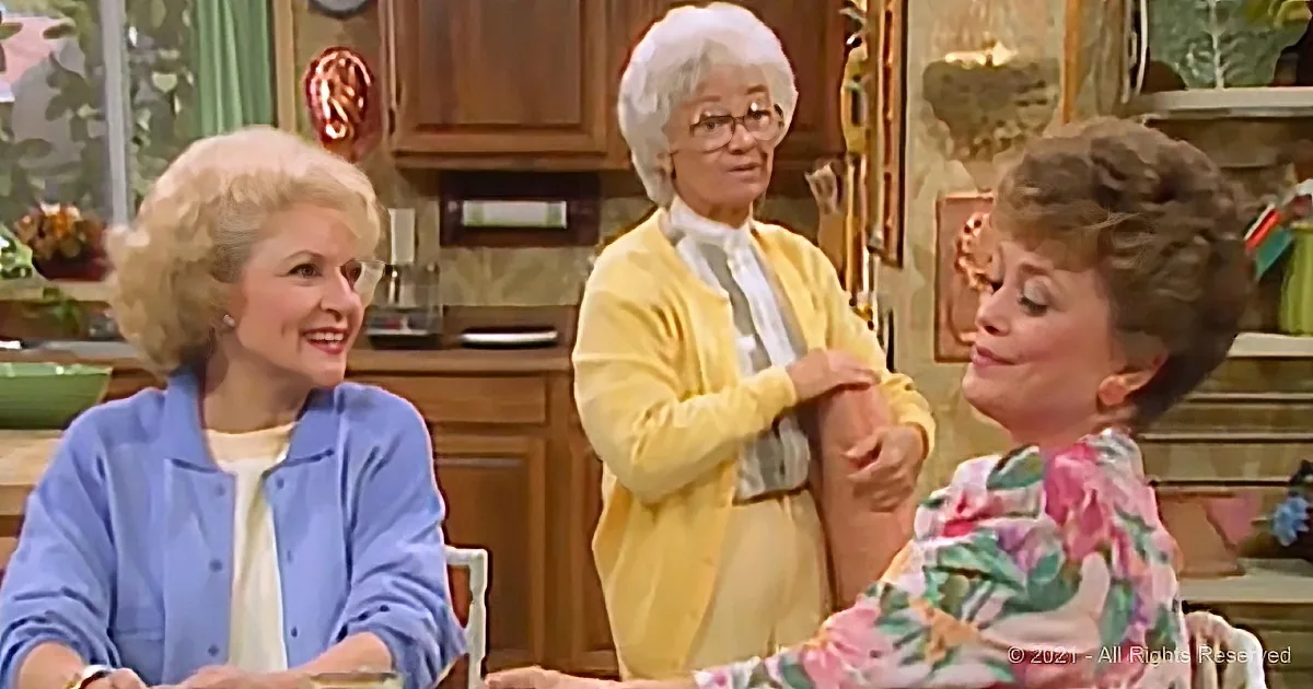 Three women in a kitchen, one in yellow cardigan adjusting her arm, another in blue smiling, third in floral shirt...