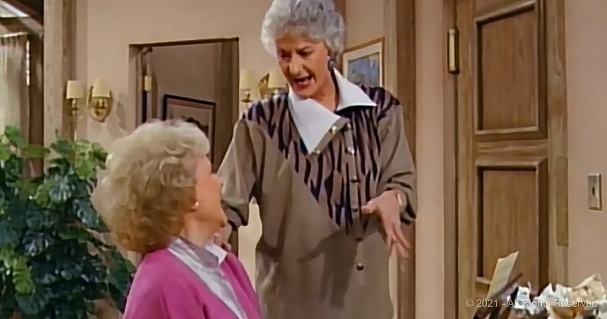 An older woman in a striped shirt is gesturing while speaking to another woman in a pink sweater in a warmly lit room.