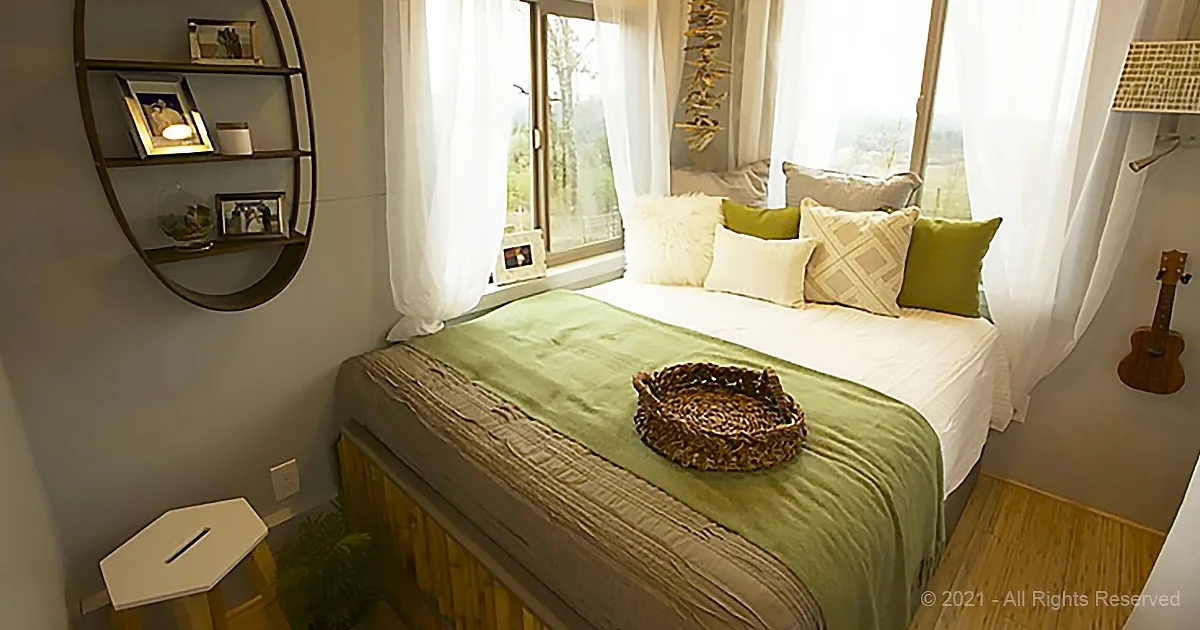 A cozy bedroom with a bed adorned in green and white, a small shelf, and a window with sheer curtains.