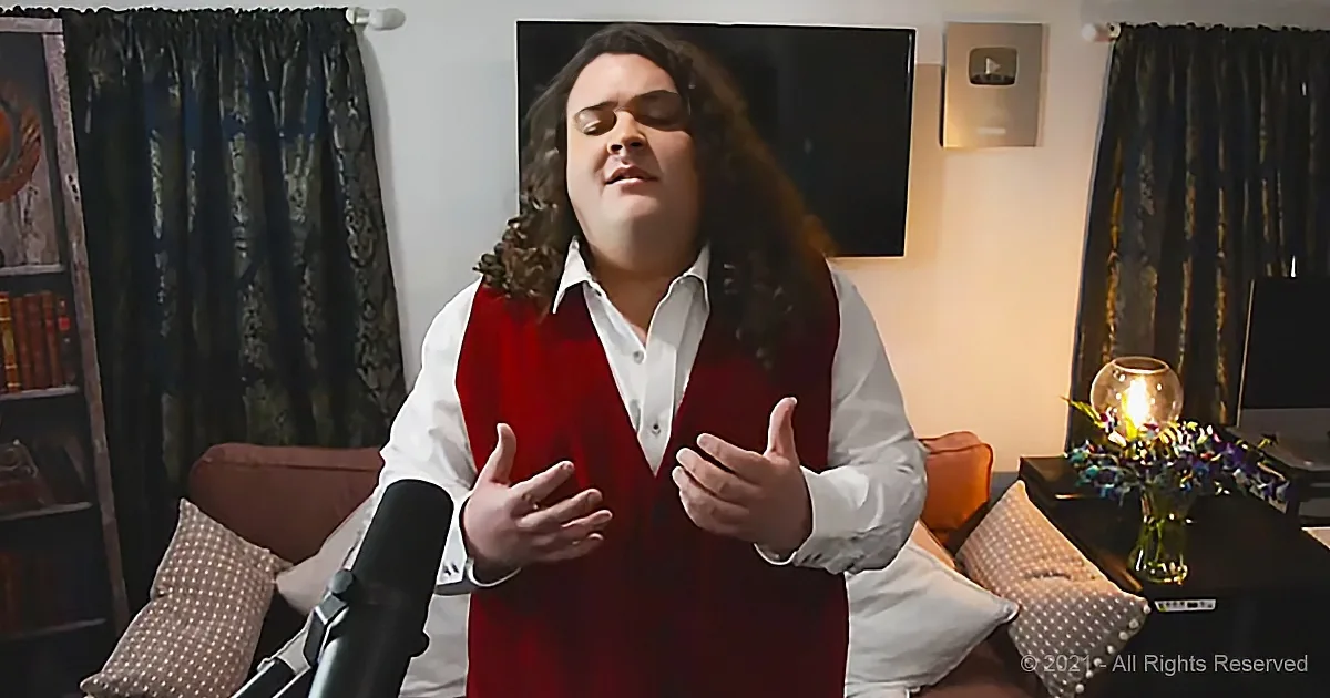A person with long hair, wearing a red vest and white shirt, gestures while speaking in a cozy room with a TV, curtains...