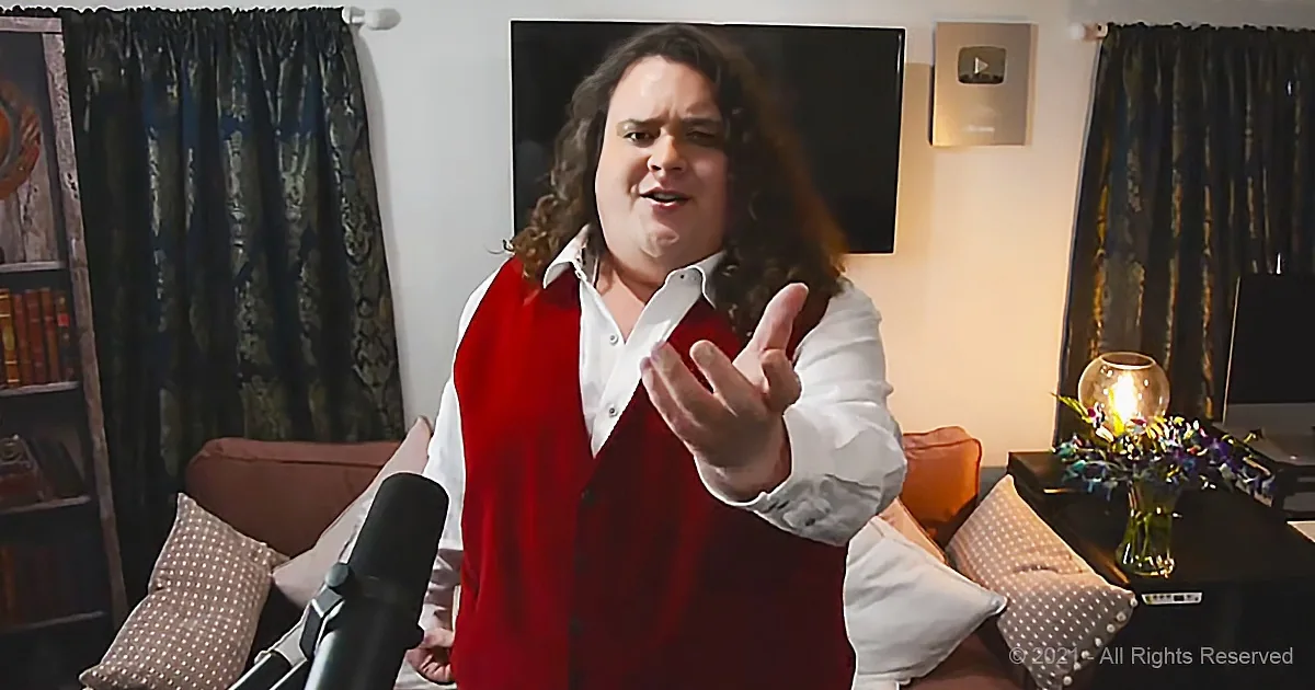 A person in a red vest and white shirt gestures while speaking in a room with curtains, a TV, and a couch.