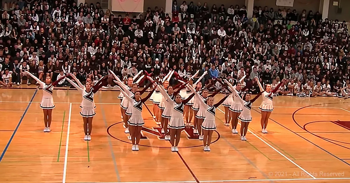 Cheerleaders in white uniforms perform on a basketball court with a large audience watching.