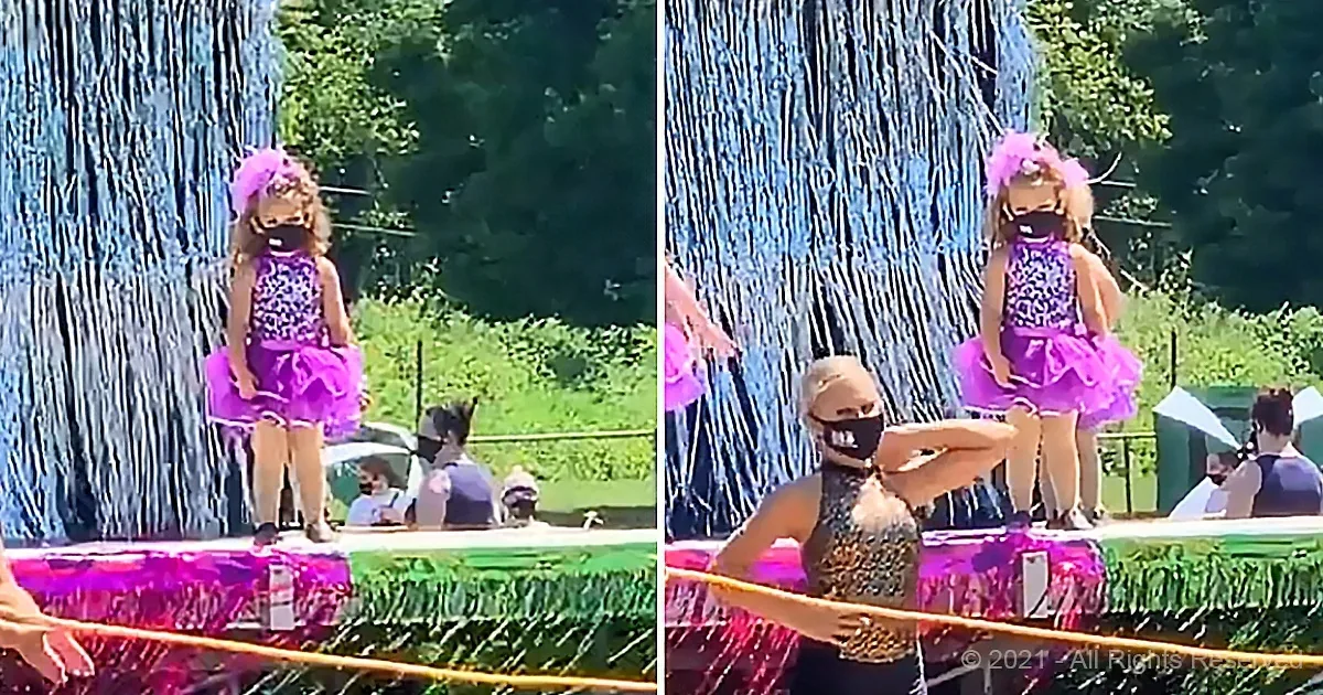 A young girl in a purple tutu stands on a float, smiling, with a woman in a black mask nearby.