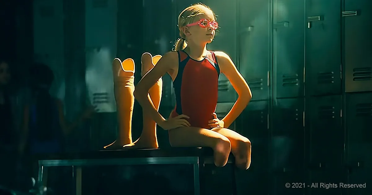 A young girl in a red swimsuit sits on a bench, wearing pink goggles, in a dimly lit locker room.