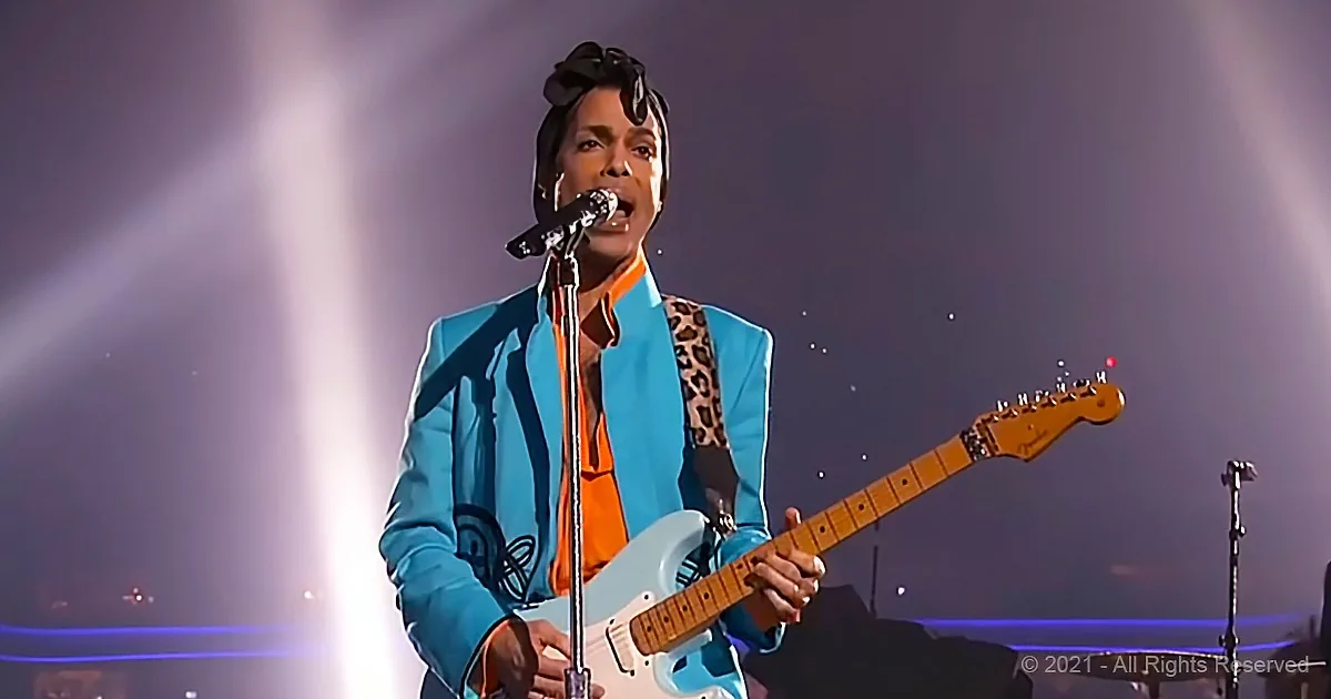 Prince in a blue suit plays a white guitar on stage with a microphone.