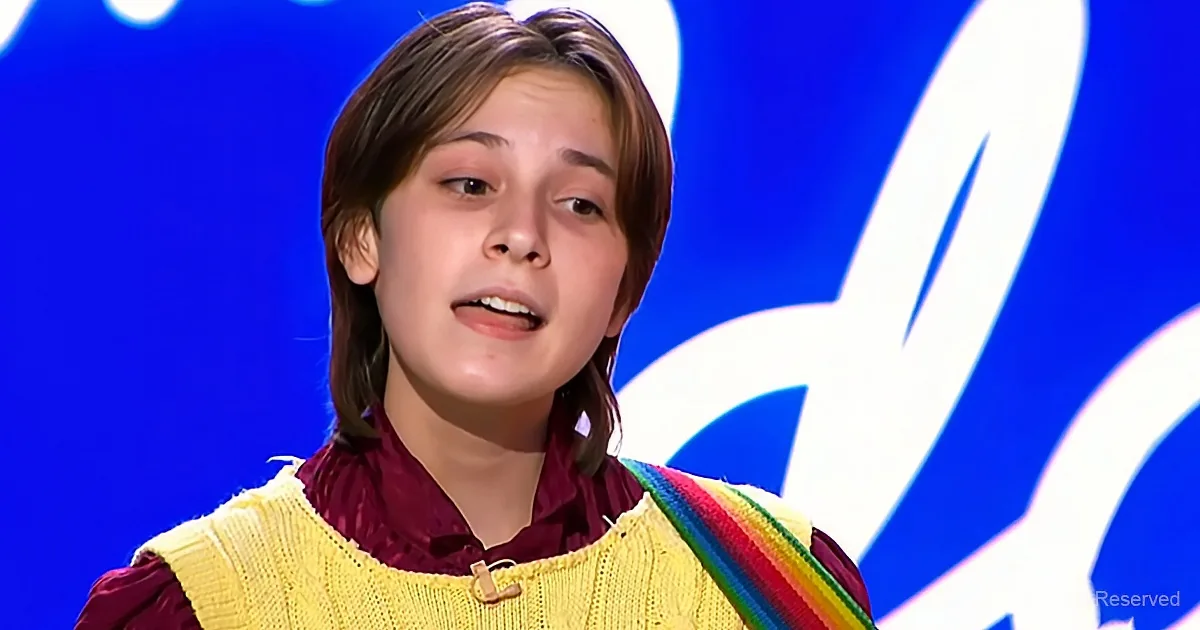 A young person wearing a yellow sweater vest and a maroon shirt, standing in front of a blue background with white...