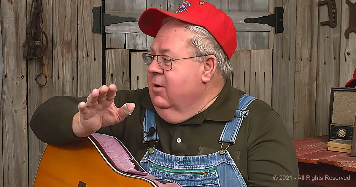 A man in a red cap and overalls gestures while holding a guitar, set against a rustic wooden backdrop.