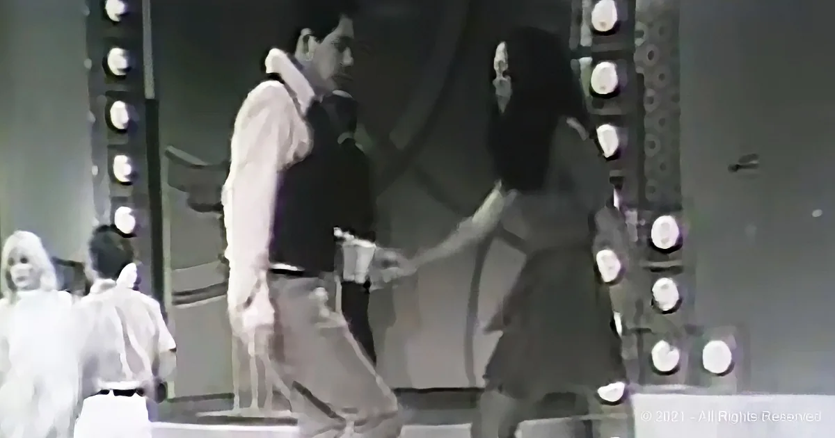 A man and woman dance on stage, surrounded by bright lights and a crowd watching.