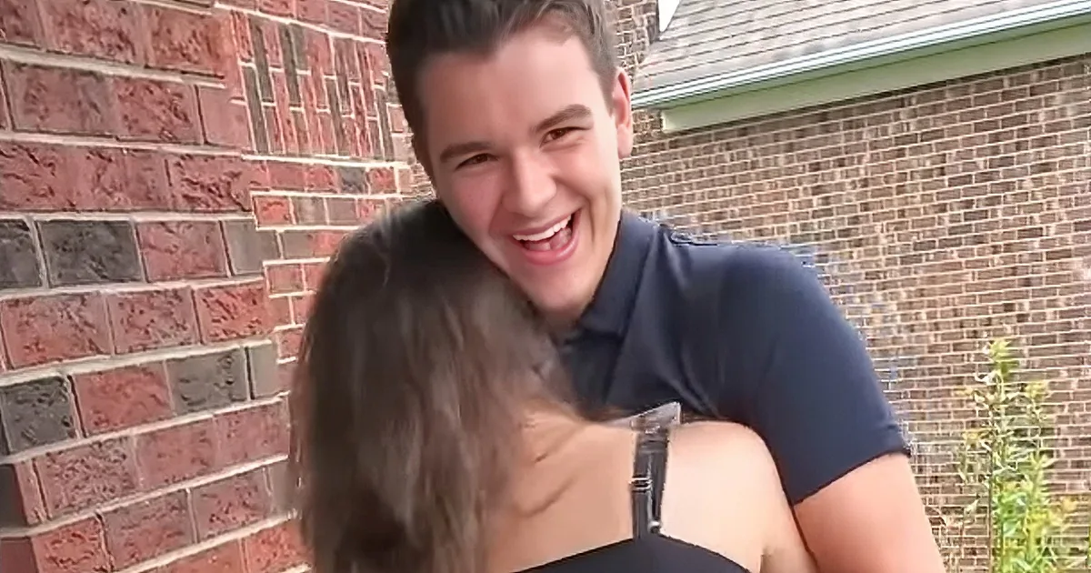 A man in a blue polo shirt is smiling and hugging a woman with long hair, both standing in front of a brick wall.