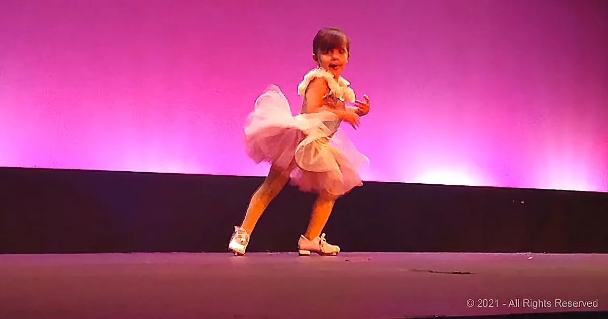A young girl in a white tutu and tap shoes dances on stage with a purple backdrop.