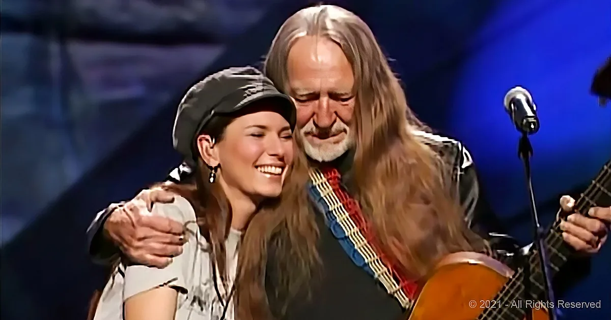 A man with long hair and a woman in a hat share a joyful moment on stage.