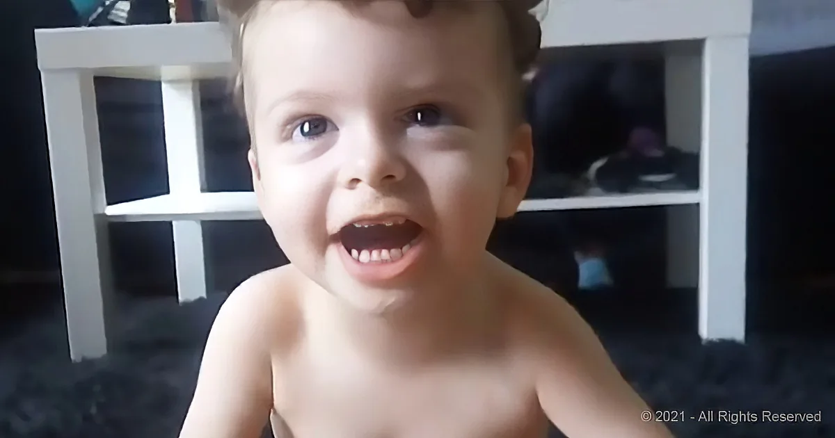 A young child with a joyful expression, shirtless, in front of a white shelf.