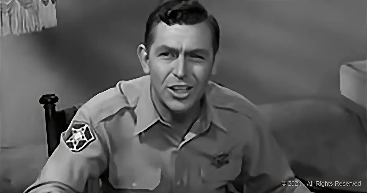 A man in a uniform with a badge on his sleeve is seated, looking slightly to the side.