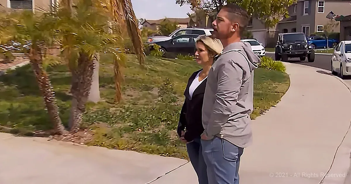 A man and woman stand on a sidewalk, looking ahead, with palm trees and parked cars in the background.