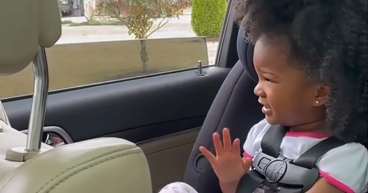 A young child in a car seat, wearing a seatbelt, appears to be speaking or reacting with an open mouth and raised hand.