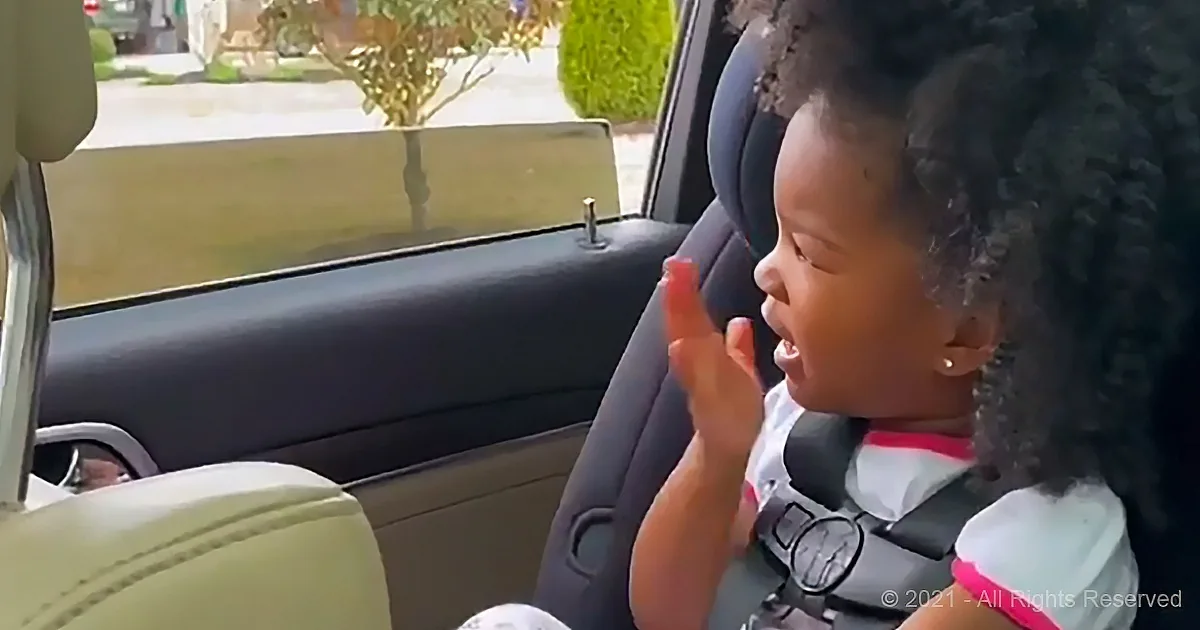 A young child in a car seat claps hands, smiling, with a suburban street visible through the window.