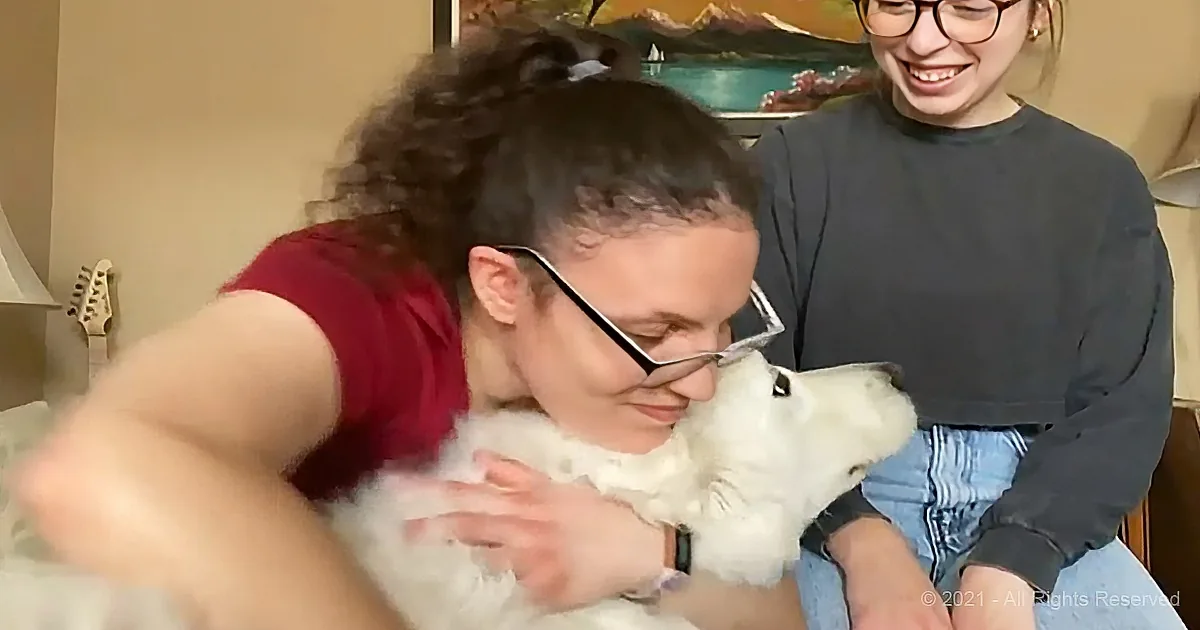 A woman in a red shirt is hugging a white dog while another woman in a black shirt smiles in the background.