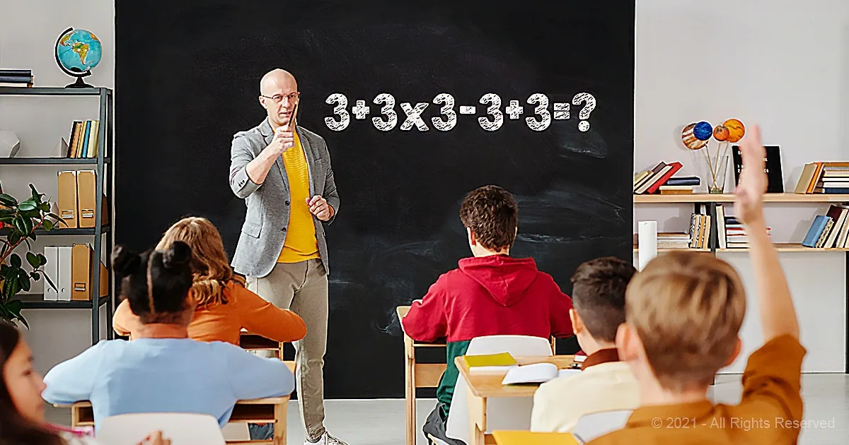 Teacher points at math problem on chalkboard while students raise hands in classroom.