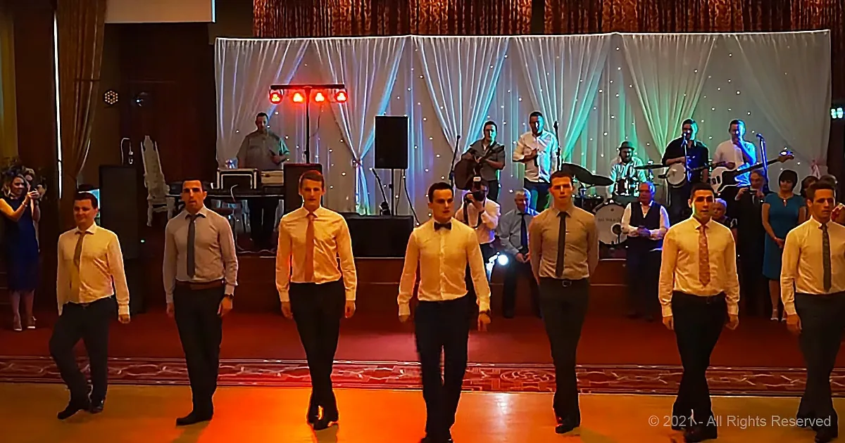 A group of men in matching outfits stands on stage, performing in front of a band and a decorated backdrop.