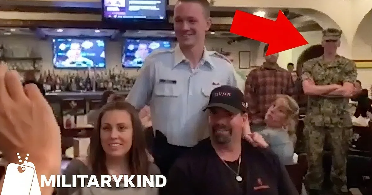 A soldier in uniform hugs another person in a bar setting with TVs and patrons in the background.