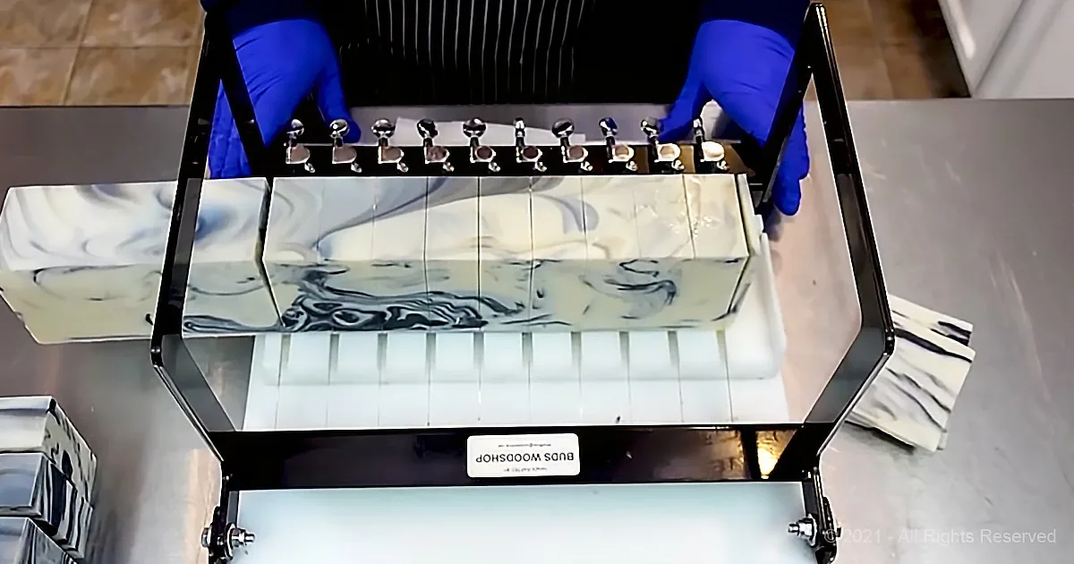 A person in blue gloves holds a tray with marbled soap bars on a metal rack.