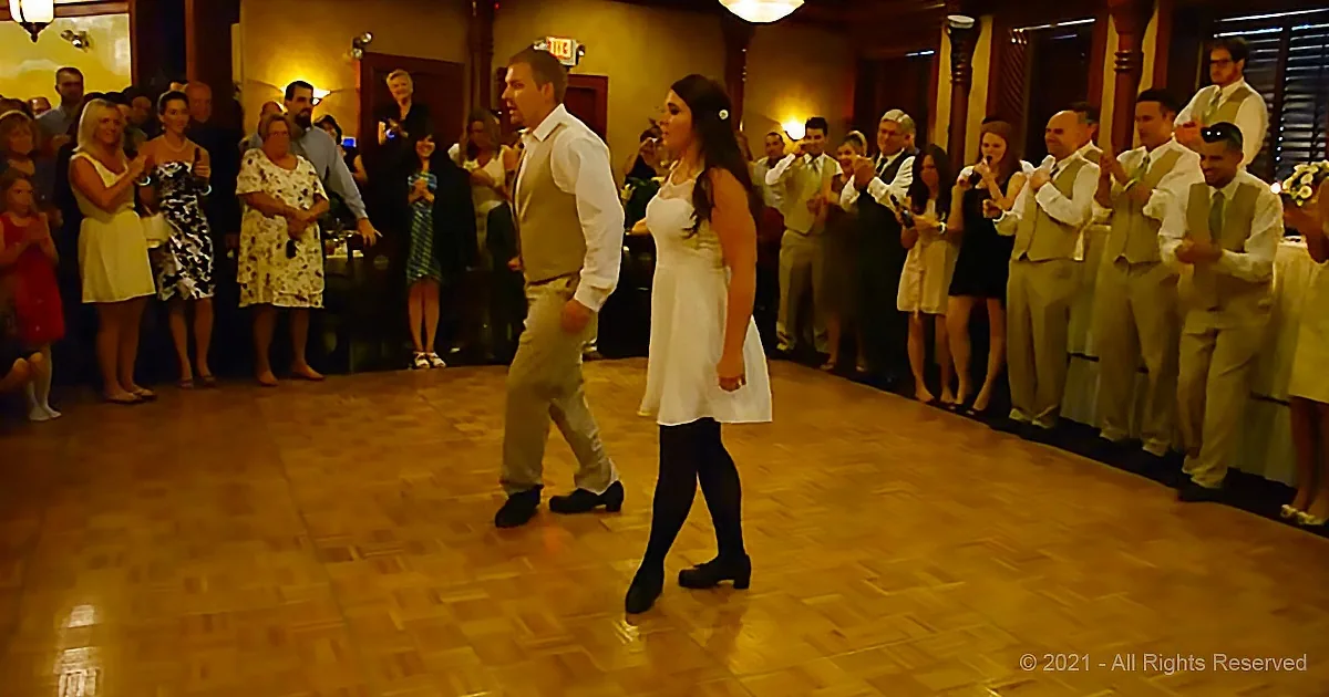 A couple dances on a wooden floor surrounded by a cheering crowd in a warmly lit room.