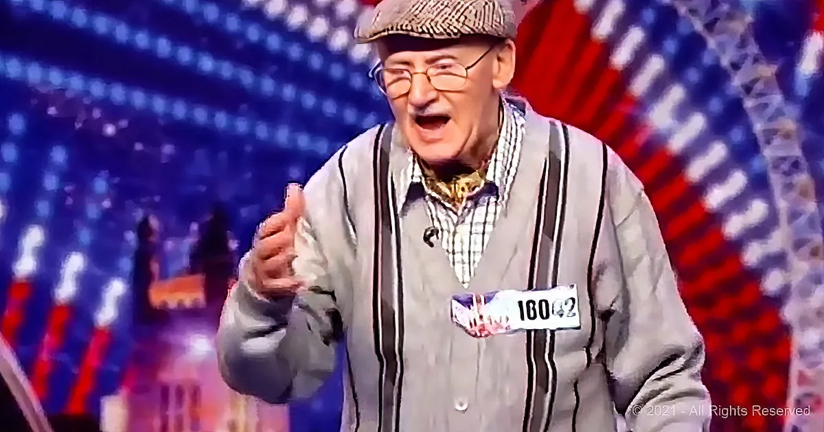 An older man in a plaid cap and striped sweater vest gestures enthusiastically on a stage with a colorful backdrop.