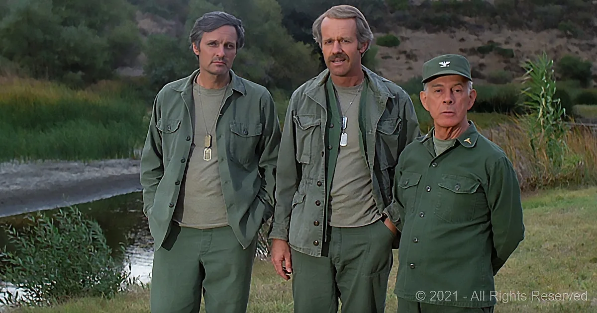 Three men in military uniforms stand outdoors, one wearing a cap, against a backdrop of grass and water.