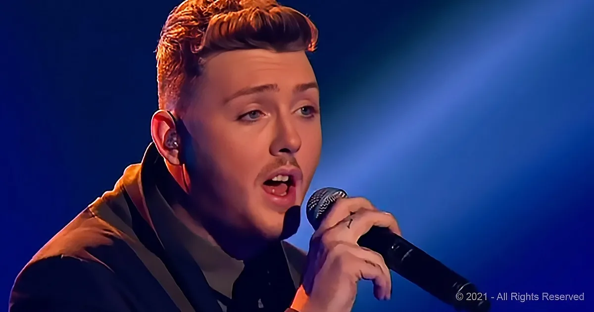 A man with short brown hair and a microphone, singing on stage with a blue light background.