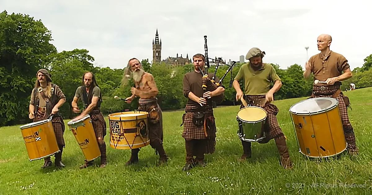 A group of musicians in traditional attire plays bagpipes and drums in a grassy field with a castle in the background.