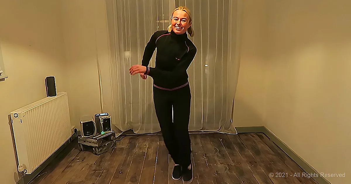A woman in a black outfit smiles while standing in a room with wooden floors, a radiator, and curtains.