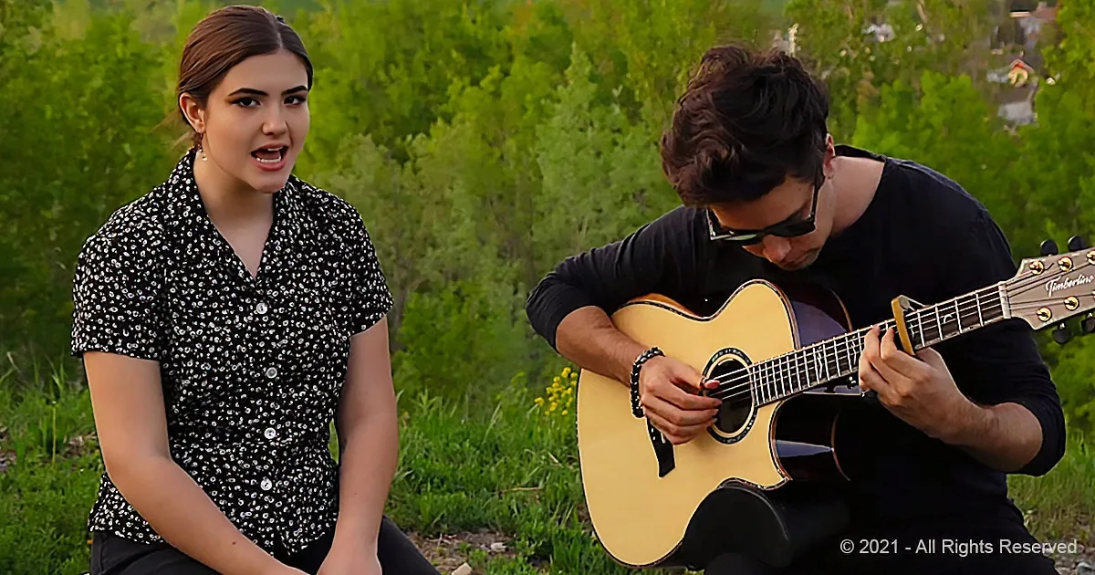 A woman sings while a man plays guitar in a lush green outdoor setting.
