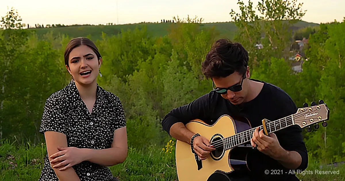 A woman sings while a man plays guitar in a lush green outdoor setting.