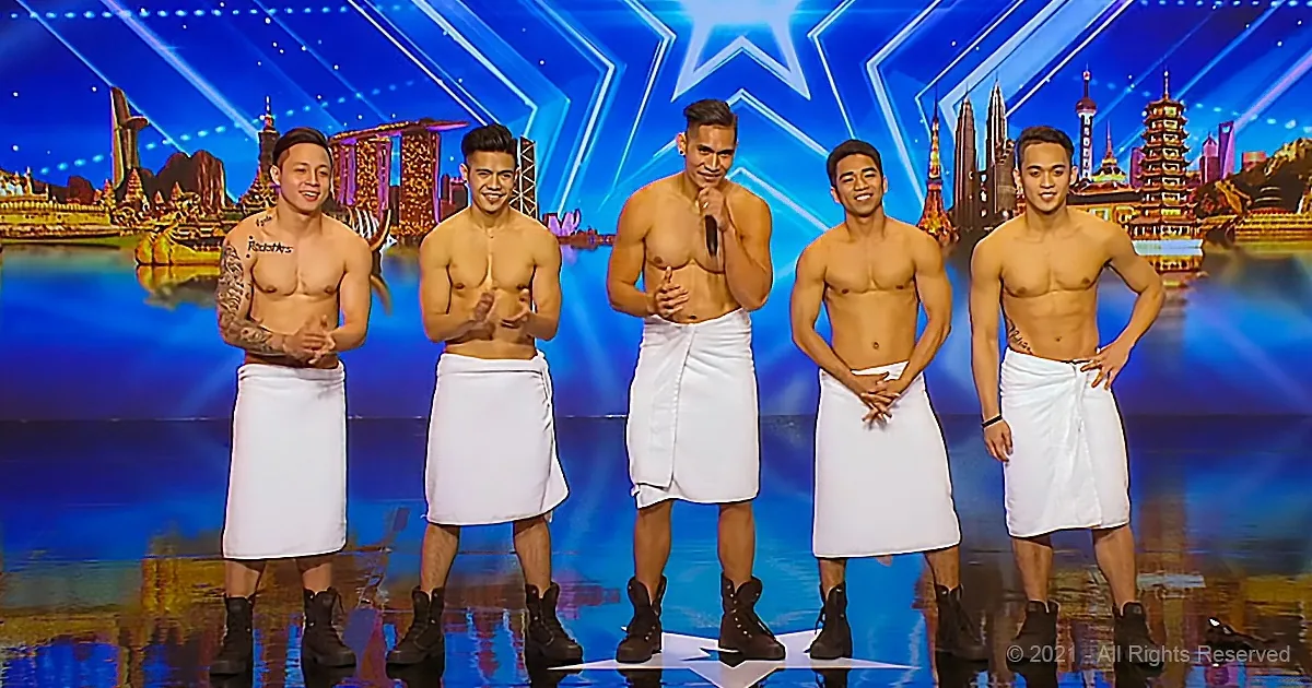 Five shirtless men in white towels and boots stand on stage, smiling, with a cityscape backdrop.