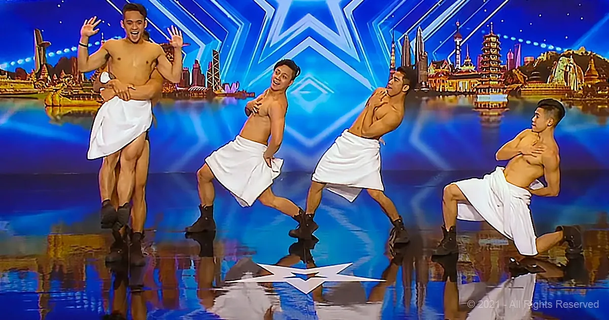 Four shirtless men in white skirts perform a synchronized dance on a stage with a cityscape backdrop.