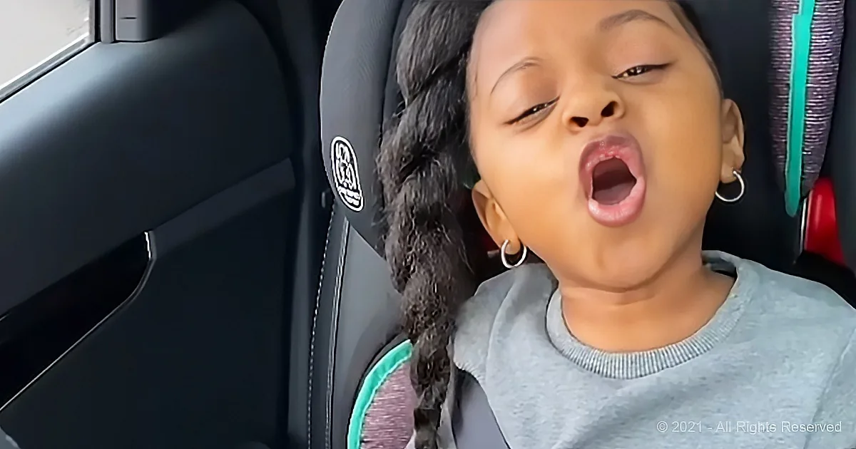 A child with braided hair sits in a car seat, mouth open, wearing a gray shirt and hoop earrings.