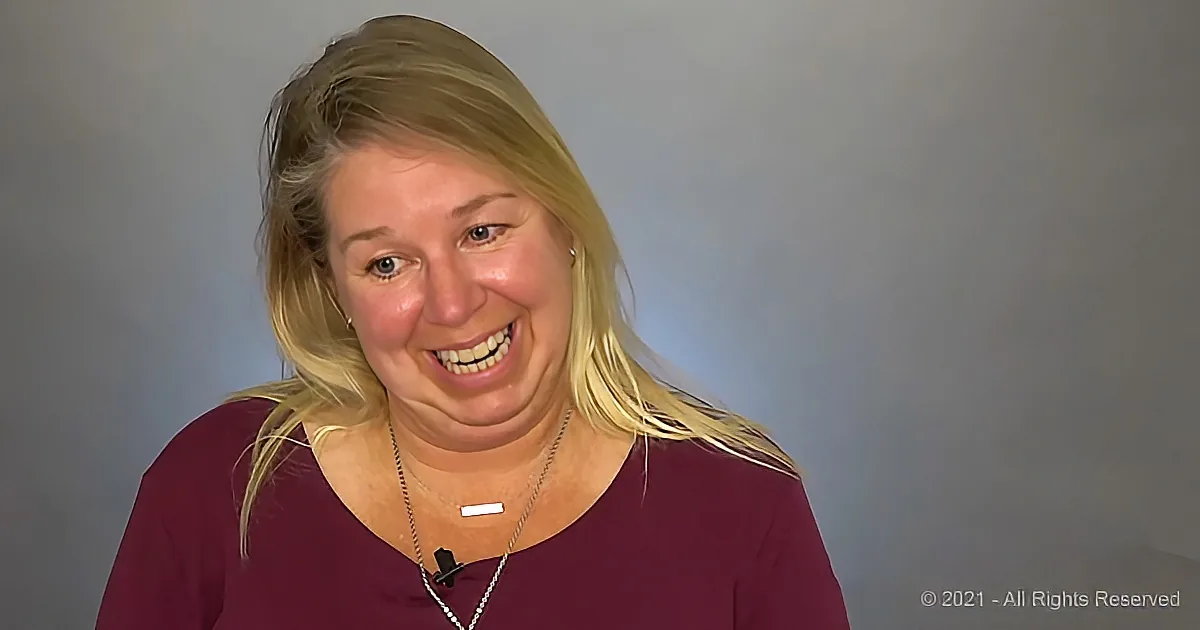 A smiling woman with blonde hair, wearing a maroon top and a necklace, stands against a plain background.