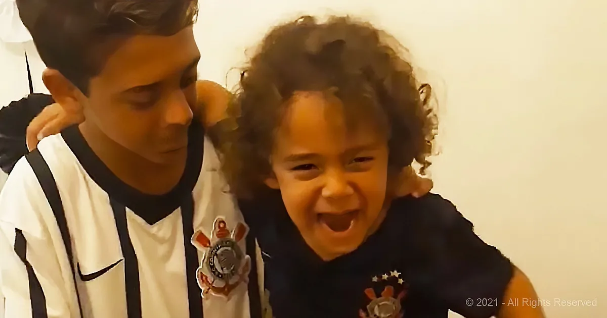 A young boy in a white shirt with a badge is embracing a child with curly hair, who is laughing.