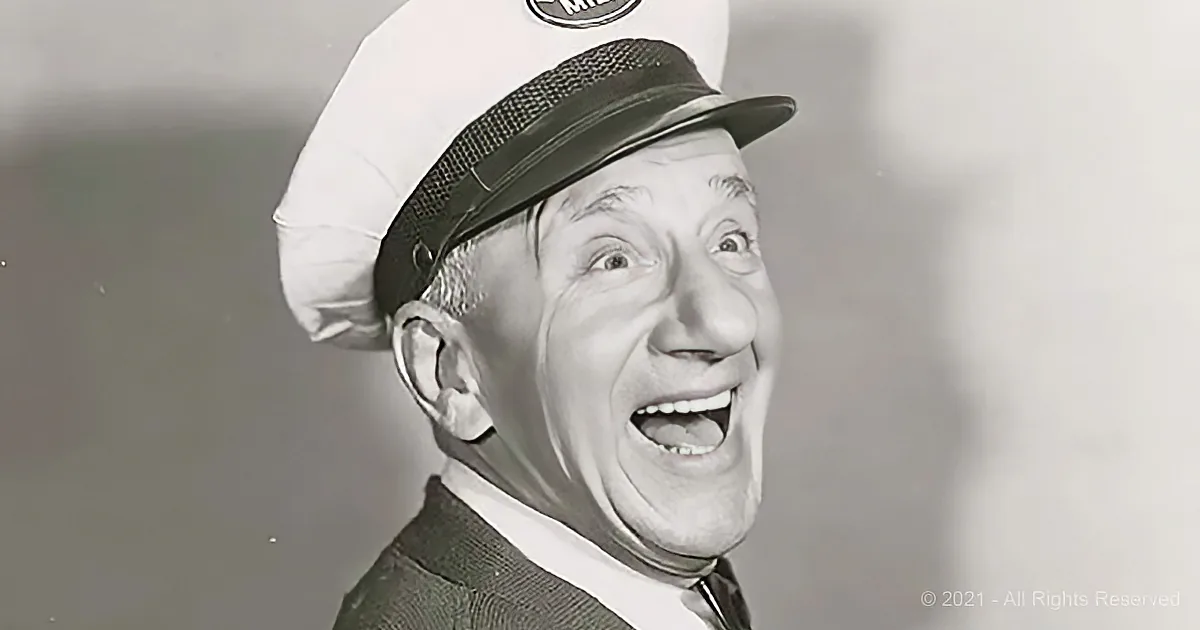 A man in a sailor hat and sweater is smiling widely, showing his teeth.
