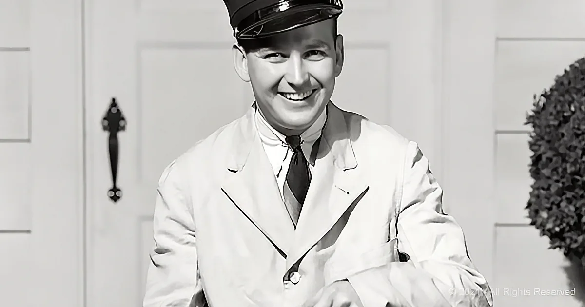 A smiling man in a white uniform and hat stands in front of a door, with a bush to the right.