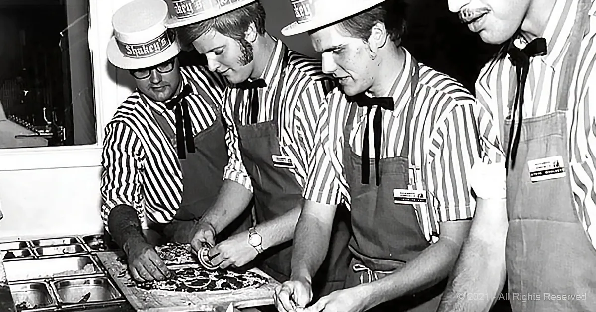 Four men in striped shirts and hats are cutting a pizza together in a kitchen.