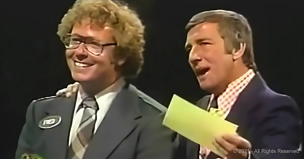 Two men in suits, one holding a yellow card, smiling and appearing to be on stage.