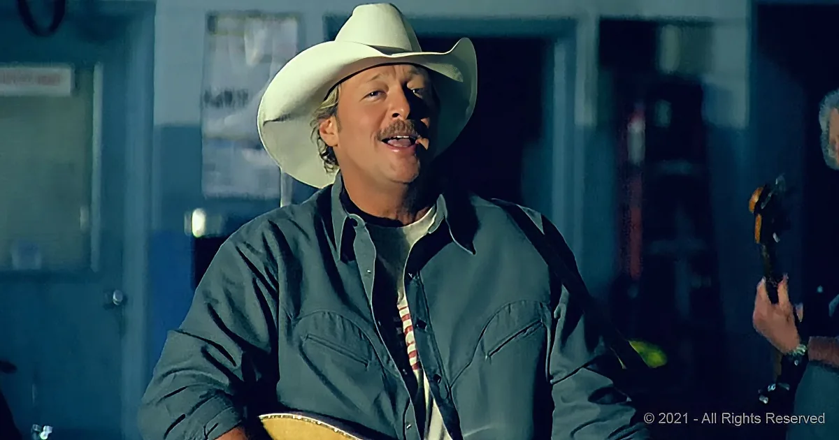 A man in a cowboy hat and green shirt sings while holding a guitar, with another person playing a guitar in the background.
