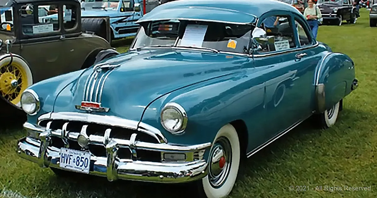 A teal vintage car with white wall tires and chrome accents is displayed at a car show.